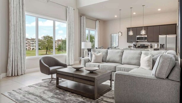 A modern living room with a sofa, coffee table, and dining area, large windows with curtains, and a kitchen in the background. - Golf View Penthouse w/ Rooftop Terrace Near Disney (Kissimmee)