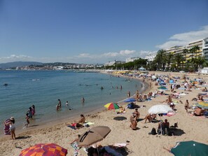 Una playa concurrida donde gente se toma el sol y nada, con sombrillas y un paisaje urbano al fondo.