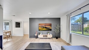 Ein modernes Wohnzimmer mit Holzboden, einem schwarzen Couchtisch, einem grauen Sofa, einem an der Wand montierten Fernseher und einem großen Fenster mit Vorhängen.