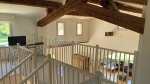 Ein geräumiges Zimmer mit hoher Decke, freiliegenden Holzbalken, einem weißen Geländer und Blick auf einen Garten durch ein Fenster.