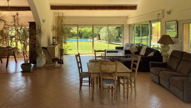 Ein geräumiger Wohnbereich mit Esstisch, Stühlen, einer Couch und einem großen Fenster mit Blick auf Garten und Pool.