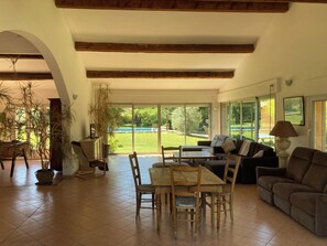 Un vaste espace de vie comprenant une table à manger, des chaises, un canapé et une grande fenêtre donnant sur un jardin et une piscine.