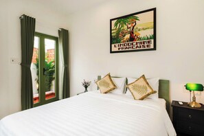 A hotel room with a bed, a nightstand with a lamp, a window with curtains, and a poster on the wall.
