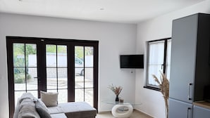 Un salon moderne avec un canapé d’angle gris, une table basse en verre, un téléviseur à écran plat fixé au mur et une grande fenêtre donnant sur l’extérieur.