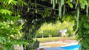 Un espace aménagé au bord de la piscine, avec une terrasse en pierre, un fauteuil de jardin en osier et une table agrémentée d’un vase de fleurs.