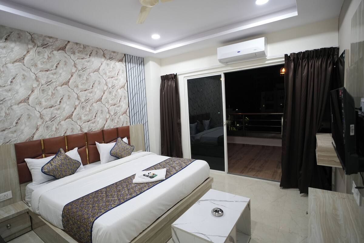 A modern hotel room with a large bed, a nightstand, a small table, a ceiling fan, and a sliding glass door leading to a balcony.