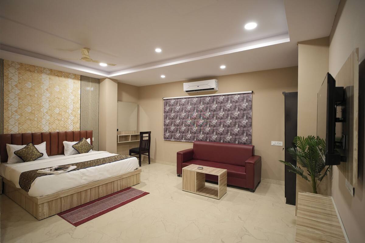 A modern hotel room with a large bed, a red sofa, a small wooden coffee table, and a potted plant.