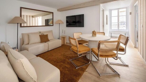A modern living room with a sofa, a coffee table, and chairs. There is a TV mounted on the wall and a window providing natural light. - NEXT TO CAFÉ DE FLORE & LES DEUX MAGOTS -3 BEDROOM PARISIAN CHARM AT RUE GOZLIN (Paris)