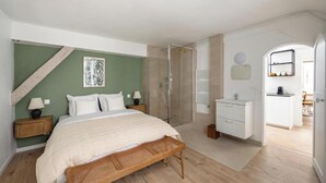 A bedroom with a bed, bedside tables, a nightstand, a wooden bench, a glass shower enclosure, a white vanity, and a small kitchen area with a sink and shelves. - NEXT TO CAFÉ DE FLORE & LES DEUX MAGOTS -3 BEDROOM PARISIAN CHARM AT RUE GOZLIN (Paris)