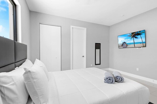 Een moderne slaapkamer met een groot bed, witte muren, een aan de muur bevestigde televisie en een schilderij van een strandgezicht.