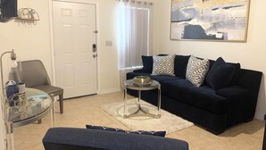 A living room with a dark blue sofa, a glass coffee table, a small side table, a chair, and a door.