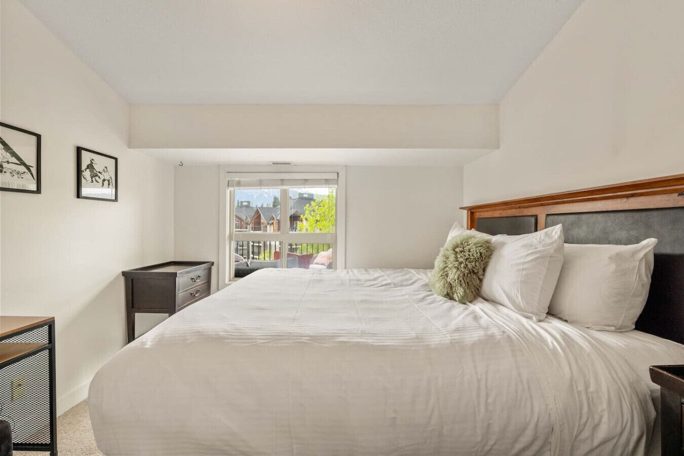 A bedroom with a large bed, a wooden headboard, a small side table, and a view of a balcony with houses in the background.