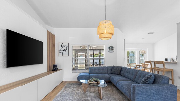 A modern living room with a large flat-screen TV, a blue sectional sofa, a glass coffee table, and a hanging rattan light fixture.