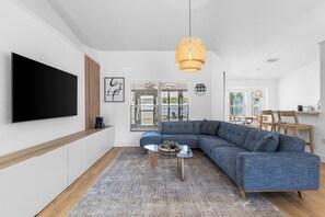 Un salon moderne doté d’un grand téléviseur à écran plat, d’un canapé d’angle bleu, d’une table basse en verre et d’un luminaire suspendu en rotin.