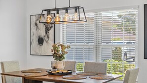 A dining area with a wooden table, grey upholstered chairs, a hanging light fixture with three bulbs, a vase with flowers, and a framed horse painting on the wall.