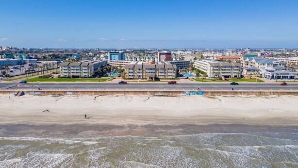 A coastal cityscape with a beachfront hotel featuring multiple-story buildings, a parking area, and a view of the ocean. - Galveston condo on Seawall near beach, gulf view, pools, WiFi, fitness room. (Galveston)