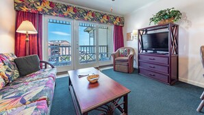 A room with a sofa, a table, a television on a dresser, and a balcony with a view. - Galveston condo on Seawall near beach, gulf view, pools, WiFi, fitness room. (Galveston)