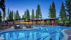 A swimming pool with lounge chairs and tables, surrounded by tall trees and a building with multiple windows. - Aloha Mountain Haus | Luxury Golf Retreat | Club Access | Billiards (Truckee)