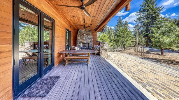 A wooden deck with a stone fireplace, a ceiling fan, and a view of the surrounding forest. - Aloha Mountain Haus | Luxury Golf Retreat | Club Access | Billiards (Truckee)