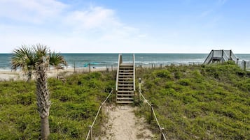 Stroll along the beach pathway to a wooden staircase, flanked by lush greenery and a palm tree, leading to a deck with ocean views.
