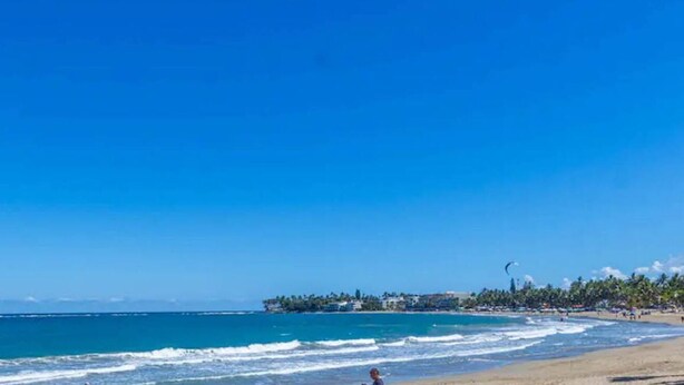 Una spiaggia con cielo sereno, mare calmo e una costa lontana con edifici e alberi.