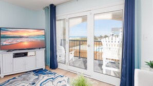 Ein Wohnzimmer mit Fernseher, einer Glasschiebetür zu einem Balkon und Blick auf den Strand.