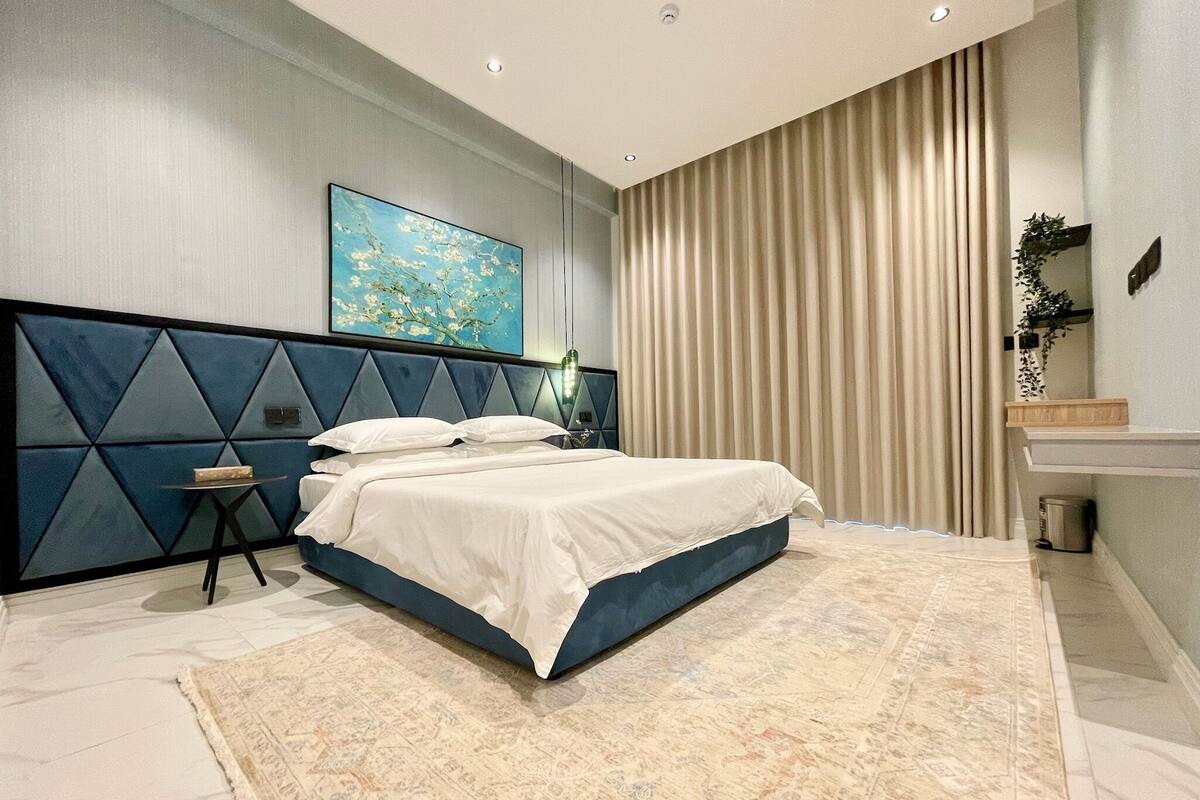 A modern bedroom with a large bed, a bedside table, a nightstand, a mirror, and a painting on the wall.