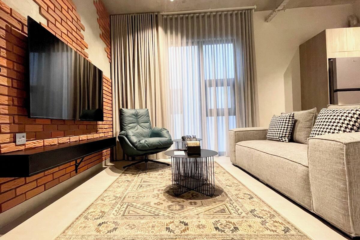 A living room with a brick accent wall, a mounted TV, a green armchair, a beige sofa, patterned rug, and a window with curtains.