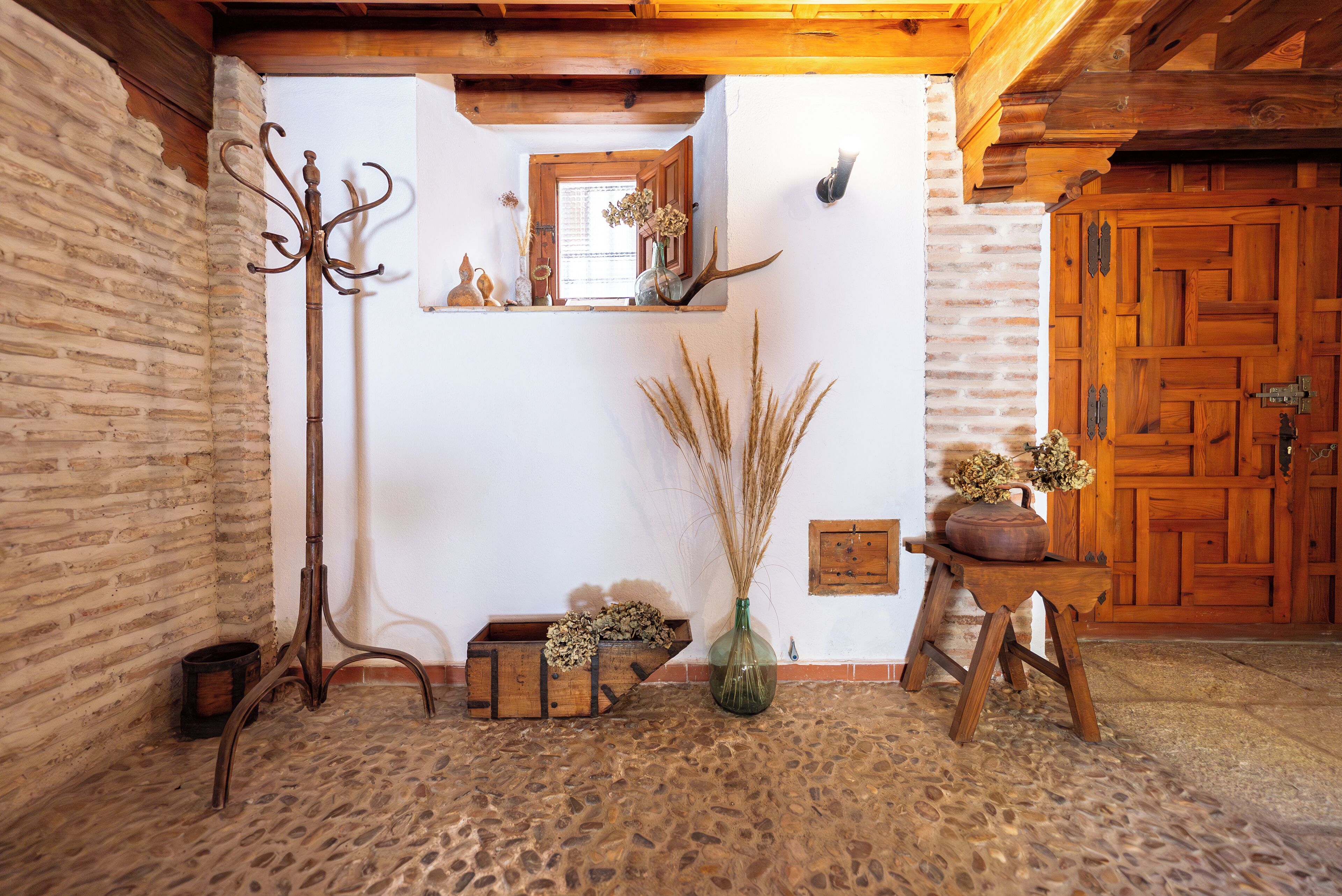 A rustic room with a stone floor, wooden beams, a coat rack, a wooden bench, a vase with dried plants, and a wooden door.