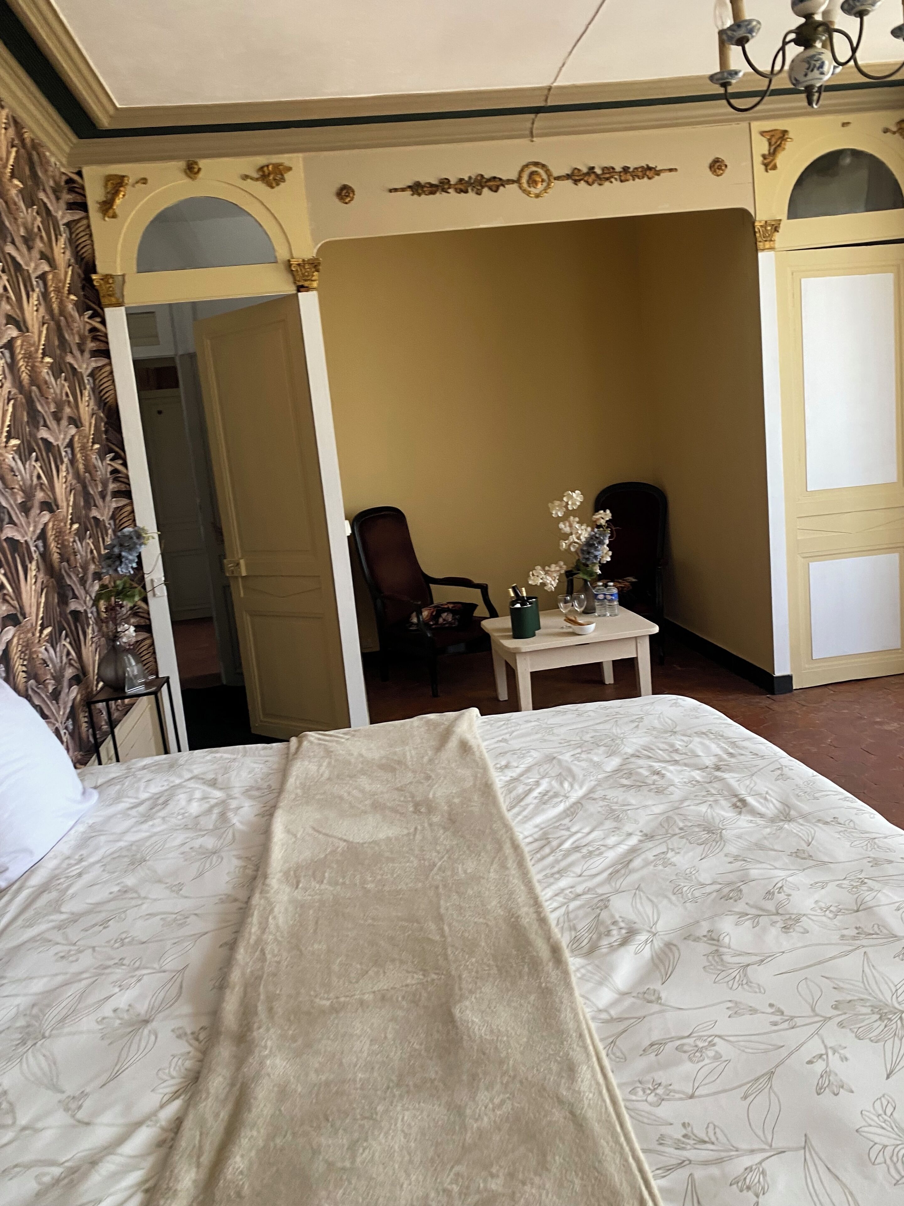 A hotel room with a bed, a small table with a vase of flowers, a chair, and a door leading to another room.