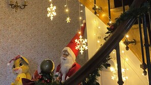 Una scala addobbata con luci a tendina e ornamenti a forma di fiocco di neve, che conduce a un pianerottolo con un muro fiorito e una statua di Babbo Natale.