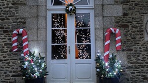 Un edificio in pietra con una porta bianca decorata da un ghirlando e bastoncini di zucchero. Davanti, due alberi di Natale illuminati.