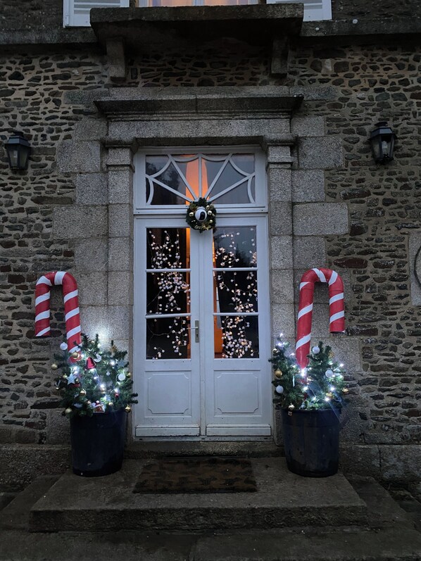 A stone building with a white door adorned with a wreath and candy canes. Two Christmas trees with lights are placed in front.