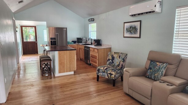 A spacious living area with a wooden floor, a beige sofa, a patterned armchair, and a kitchen with a central island.