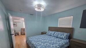 A bedroom with a bed, wooden floor, a ceiling light fixture, a window with blinds, and a door leading to another room.
