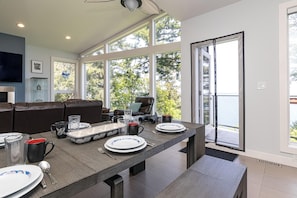 Un comedor moderno con una mesa larga dispuesta para cuatro comensales, una zona de estar acogedora con un sofá, y grandes ventanales que ofrecen vistas al exterior.