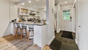 Une cuisine moderne avec des meubles blancs, un îlot central doté de tabourets de bar et une vue sur l’extérieur par une porte.