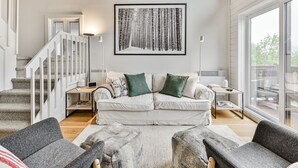 A modern living room with a white sofa, two green cushions, a grey armchair, and a coffee table. A staircase with white railings leads to another level. A large framed artwork hangs on the wall, and a window provides a view of the outdoors.