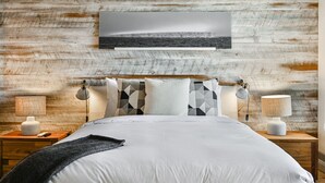 A bedroom with a wooden headboard, a large bed with white bedding, geometric patterned pillows, two bedside lamps, and a framed photograph on the wall.
