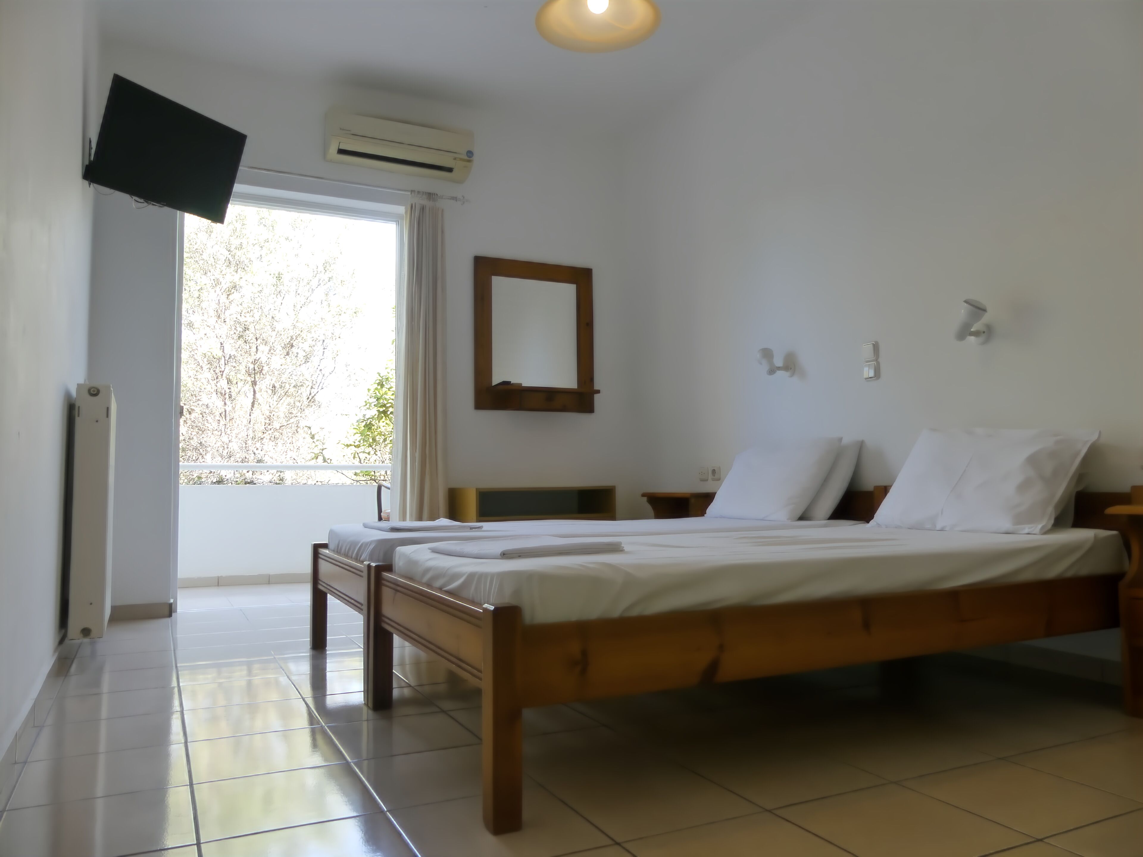 A bedroom with a wooden bed, a flat-screen TV mounted on the wall, an air conditioning unit, and a window with curtains.