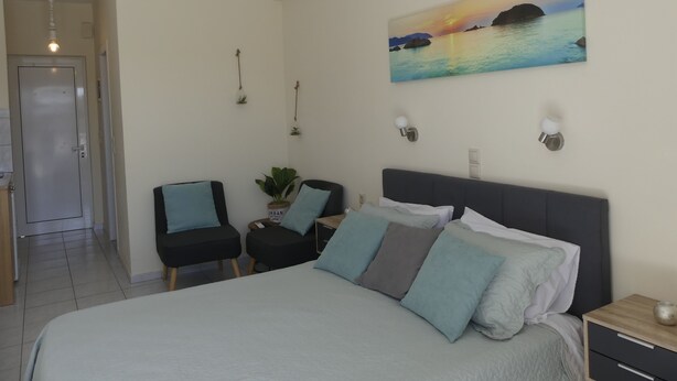 A bedroom with a bed, two chairs, a nightstand, a painting of a sunset, and a door. - AMZ Apartments (Kato Gouves)