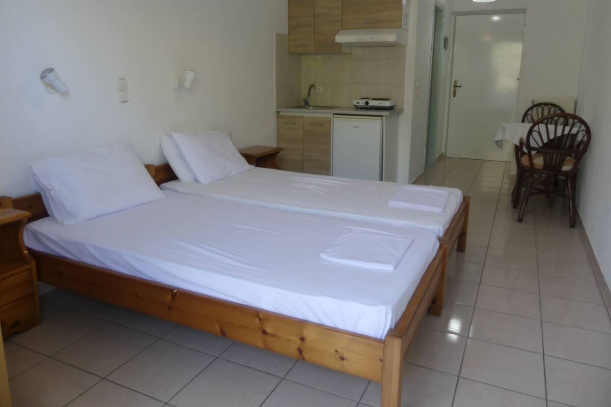 A double bed with white linens in a room with a kitchenette and a dining area.