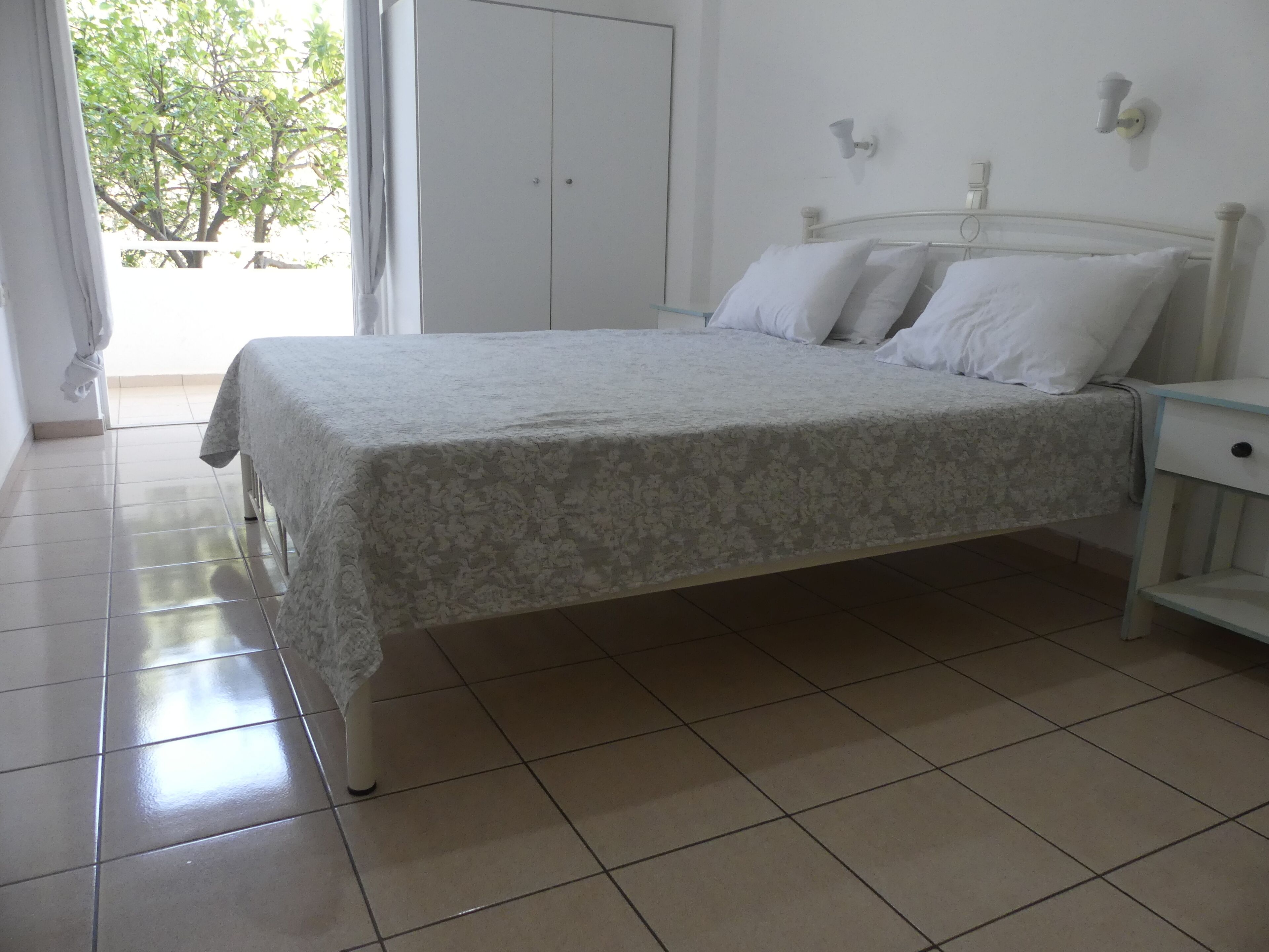 Un lit bien fait, recouvert d’une couvre-lit à motifs floraux, dans une pièce aux murs blancs et au sol carrelé.