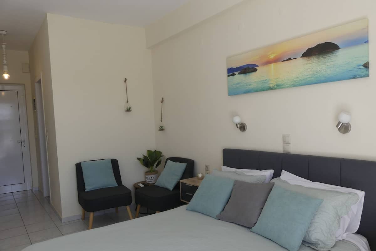 A bedroom with a bed, two chairs, a small table, a potted plant, and a painting of a sunset over water.