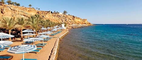 A beach with sun loungers and umbrellas, clear blue water, and a rocky cliff.