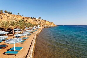 Ein Strand mit Liegestühlen und Sonnenschirmen, klares blaues Wasser und eine felsige Klippe.