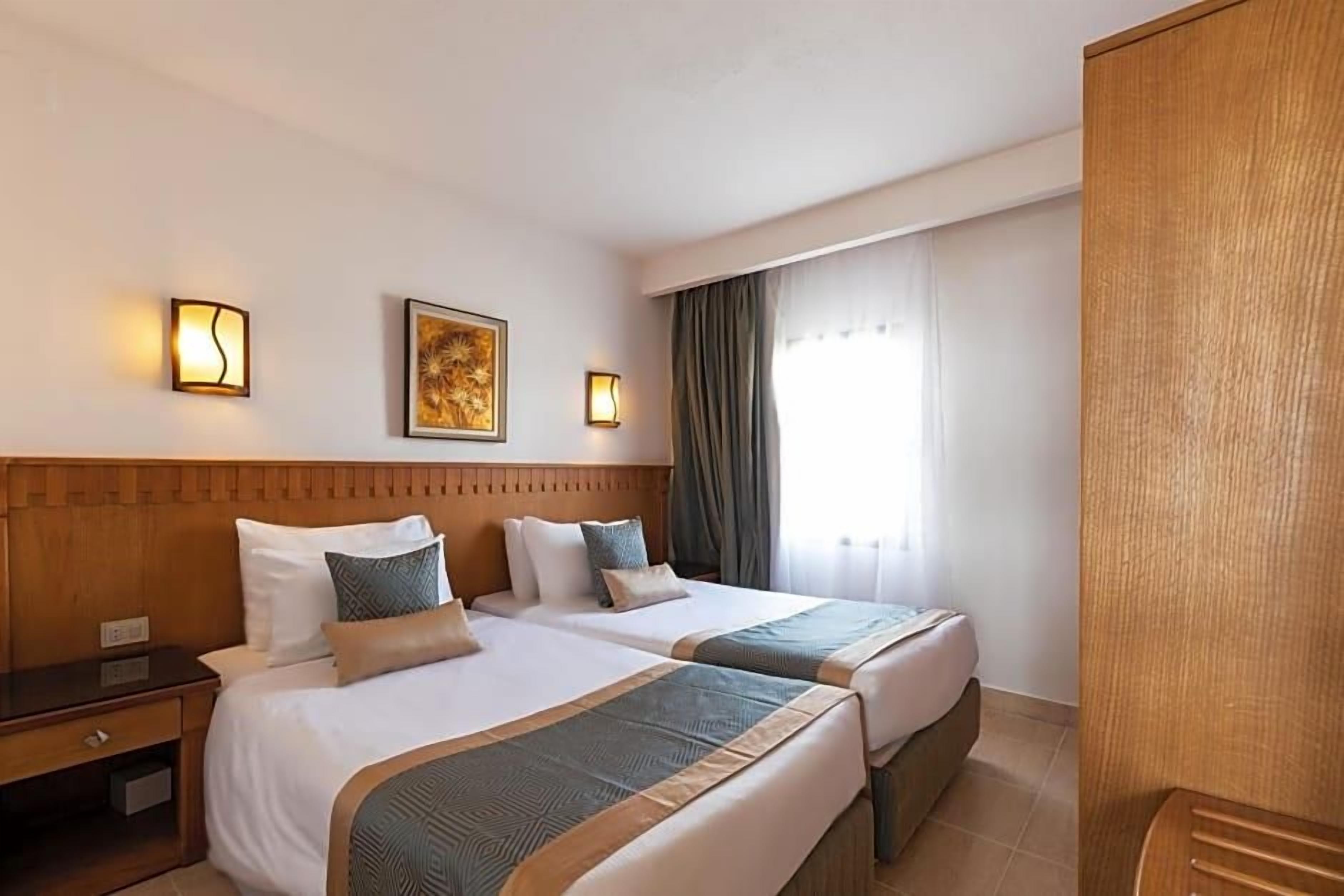 A hotel room with two beds, a wooden headboard, bedside tables, wall-mounted lights, a framed picture, and a window with curtains.