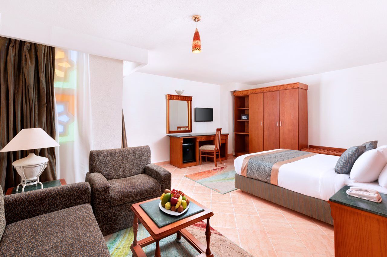 A hotel room with a bed, a desk, a chair, a sofa, and a table with fruit.