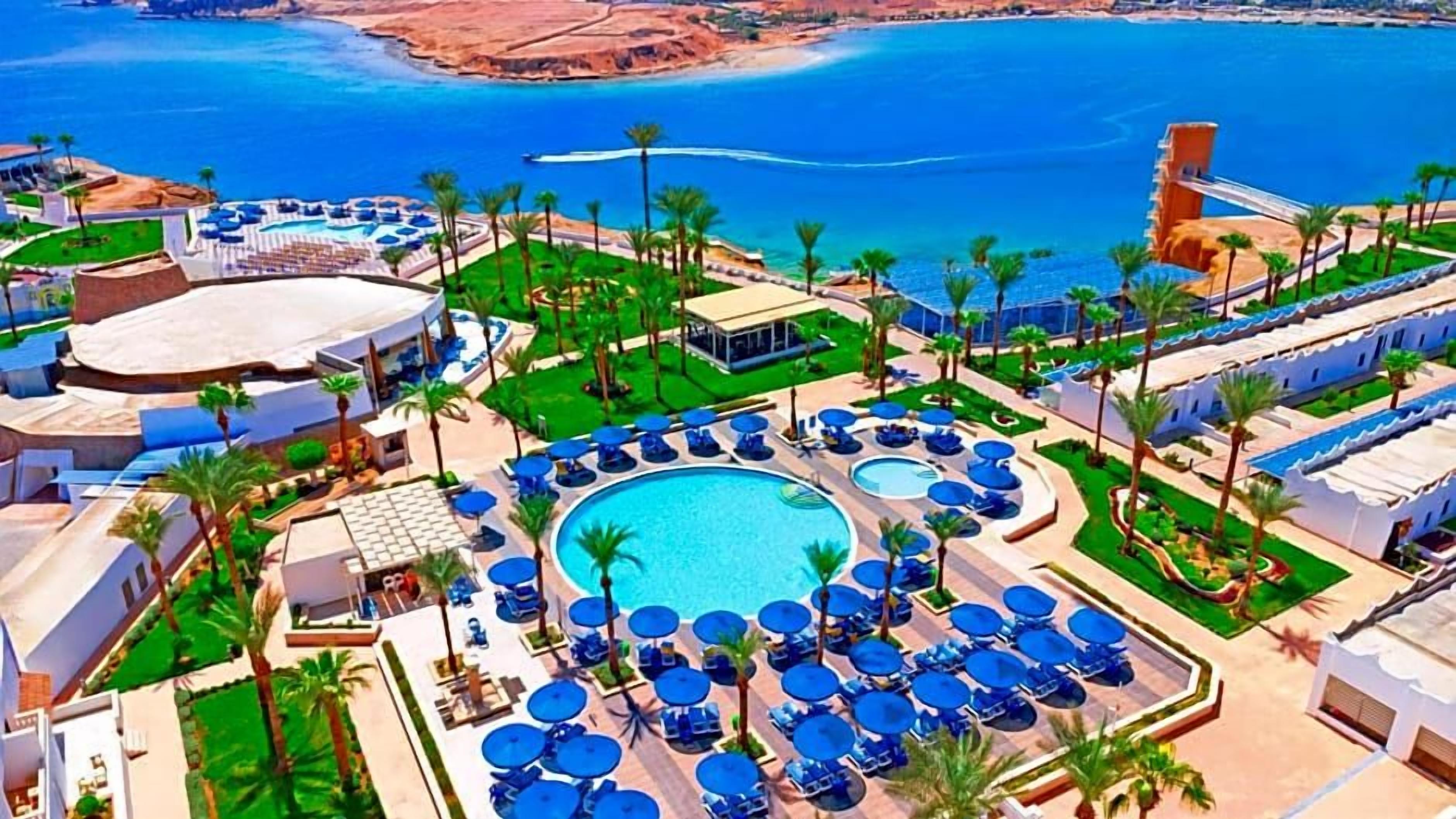 Un complexe hôtelier doté d’une piscine centrale, entourée d’espaces de détente avec des parasols, des palmiers et une vue sur la mer.