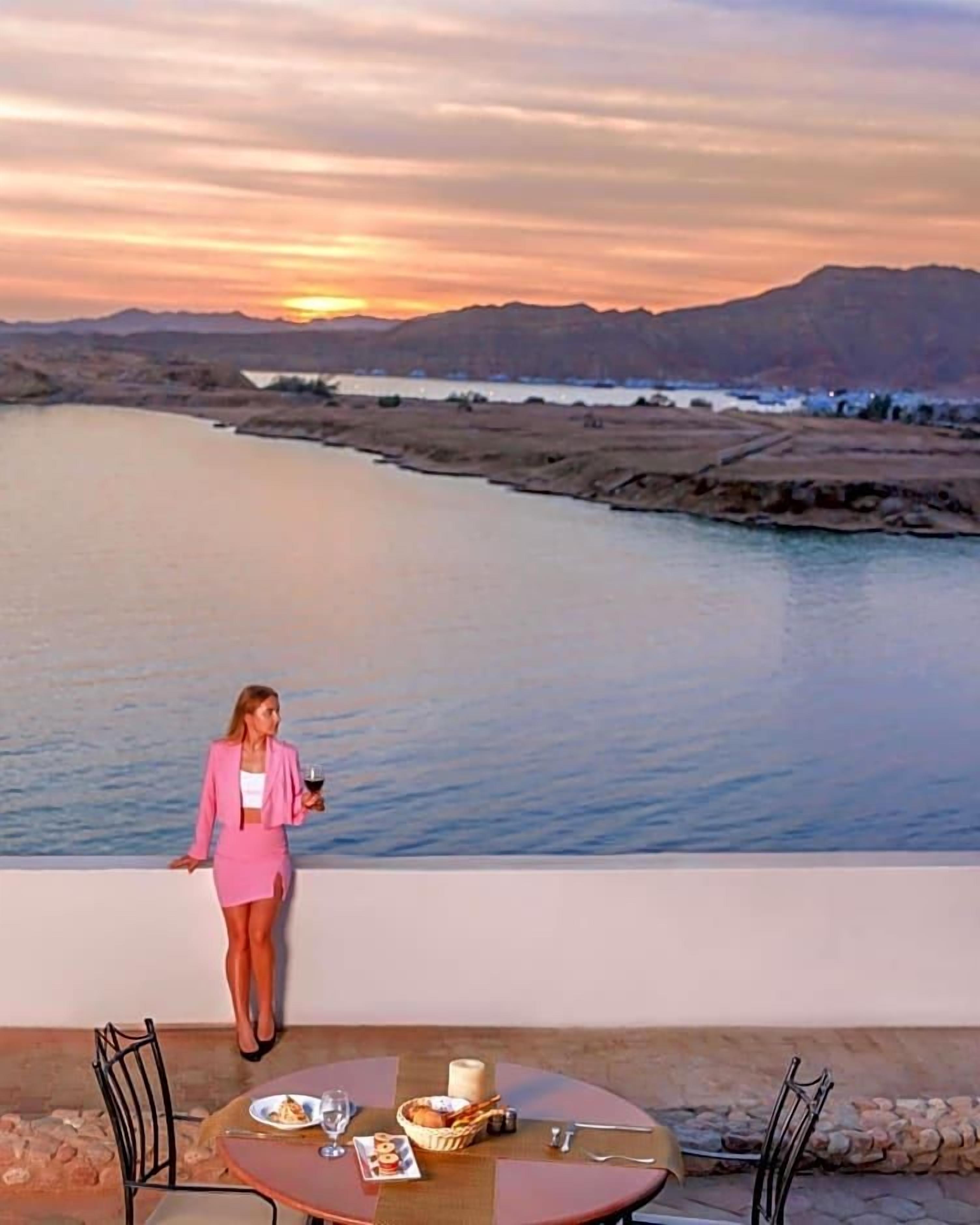 A woman in a pink suit standing by a table with a view of a river and mountains in the background.